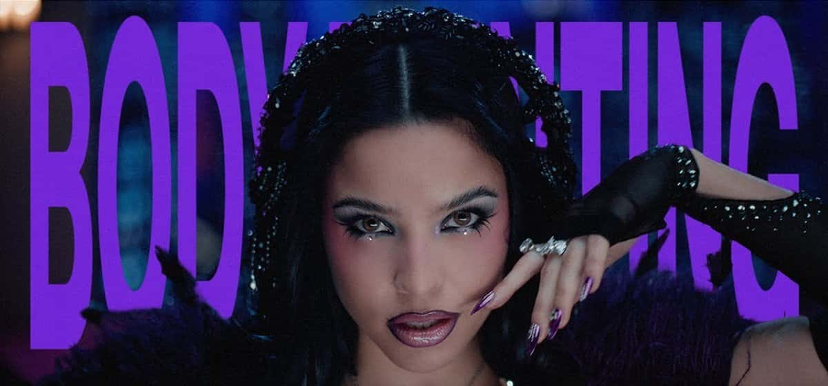 Emilia, de pelo negro con un tocado de brillos y guantes negros, mira a cámara con una mano junto a la cara, delante de la palabra «BODYPAINTING» en letras violetas, en una imagen del video oficial