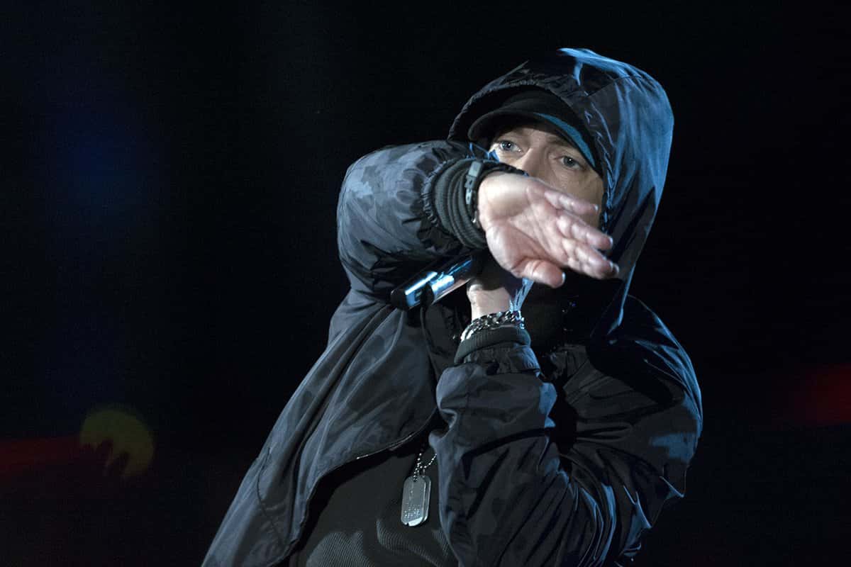 Eminem, con capucha negra, gorra y una chapa colgada del cuello, canta con el micrófono en la mano y el brazo cruzado delante de la cara, sobre fondo oscuro (foto de archivo, noviembre de 2014)