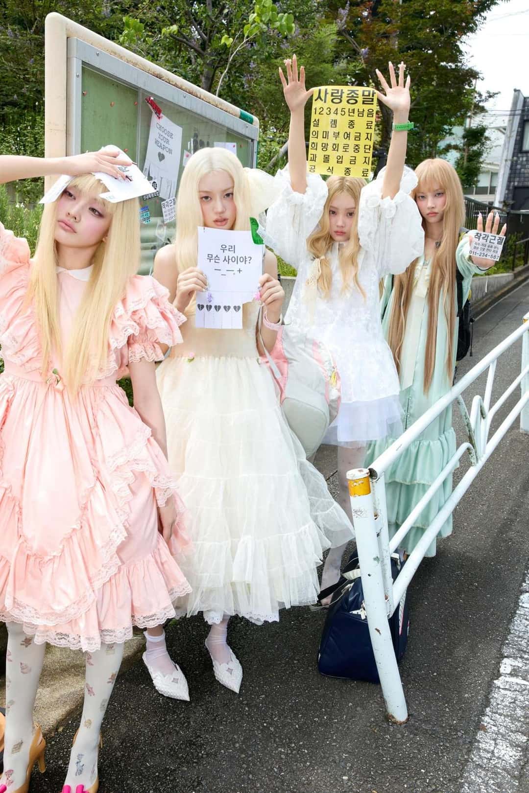 Foto conceptual de «BREAK EVEN»: las integrantes de ILLIT, con pelucas rubias y vestidos de volados en tonos pastel, posan en una calle junto a una cartelera, con carteles escritos a mano en coreano. Foto: Belift Lab