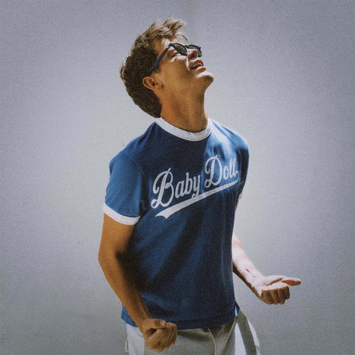 Portada del single «Babydoll», de Jamie Miller: el cantante, con anteojos de sol y una remera azul con la palabra «Baby Doll», mira hacia arriba y sonríe sobre un fondo gris
