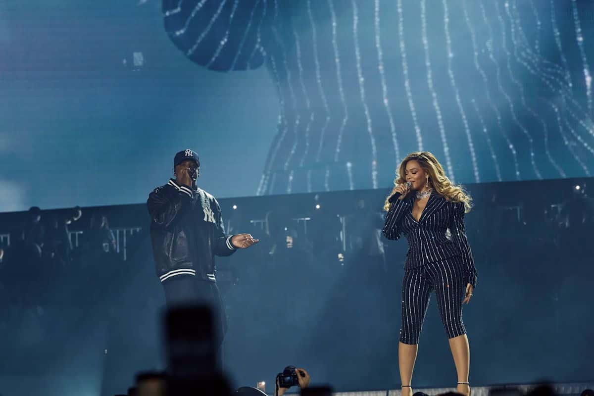 Foto oficial del primer show en el Yankee Stadium: Jay-Z, con gorra y campera de los Yankees, canta junto a Beyoncé, que lleva un traje oscuro a rayas con brillos, frente a una pantalla gigante en tonos azules