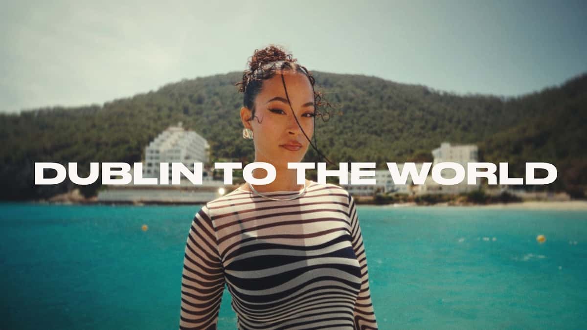 Fotograma del mini documental oficial "Dublin to the World": Jazzy con remera rayada blanca y negra, frente a un mar turquesa y una costa con hoteles blancos, con el título del film sobreimpreso