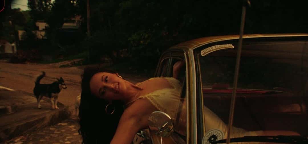 Fotograma de archivo del video oficial de «Mexico Honey», de su disco «Middle of Nowhere»: Kacey Musgraves, con vestido claro, se recuesta contra un auto viejo color dorado en una calle de tierra, con un perro husky al costado