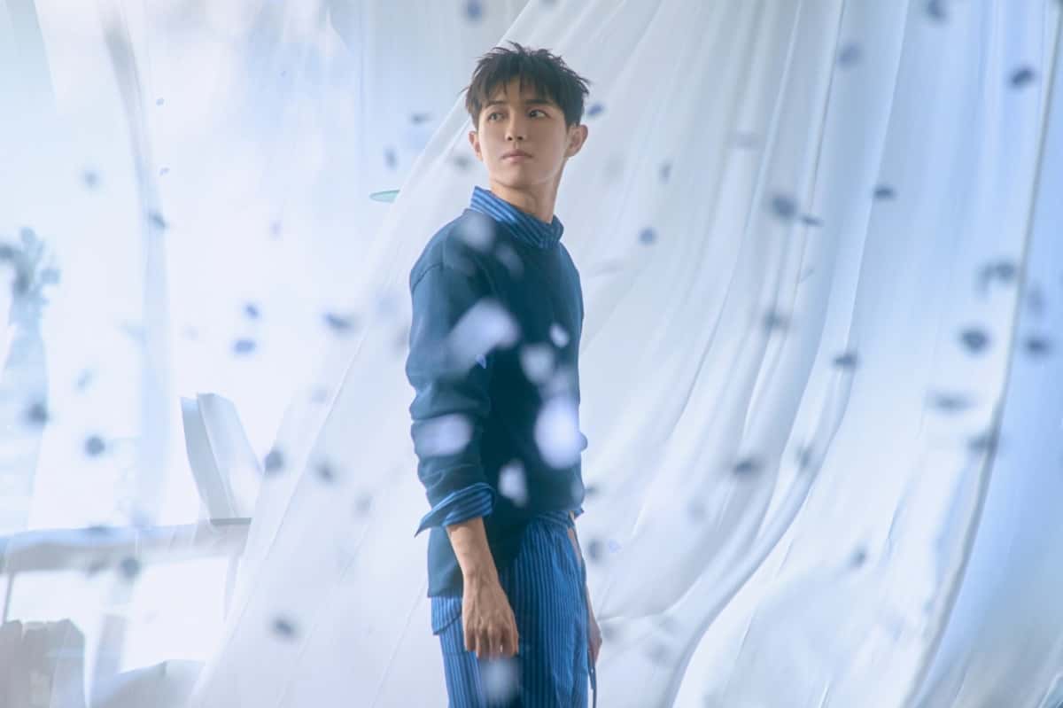 Foto conceptual oficial de Kim Jae Hwan para "Love, From Here": de pie entre cortinas blancas, con suéter azul y camisa a rayas, mirando de costado. Foto: WAKEONE