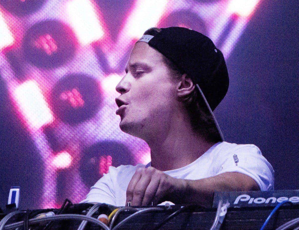 Kygo, con gorra negra hacia atrás y remera blanca, pasa música en una consola frente a una pantalla con luces violetas (foto de archivo, julio de 2016)