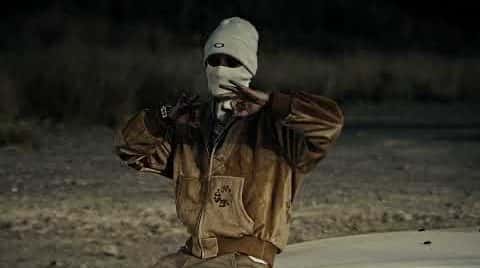 Fotograma del video oficial de «DOGGY»: una persona con pasamontañas y campera marrón hace un gesto con las manos frente a un auto blanco, de noche