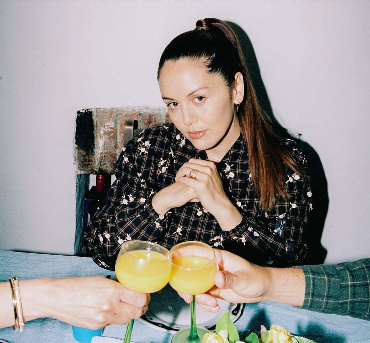 Foto de prensa de Logic1000 sentada a una mesa, con camisa a cuadros con flores bordadas, mientras dos manos brindan con copas de jugo naranja delante de ella
