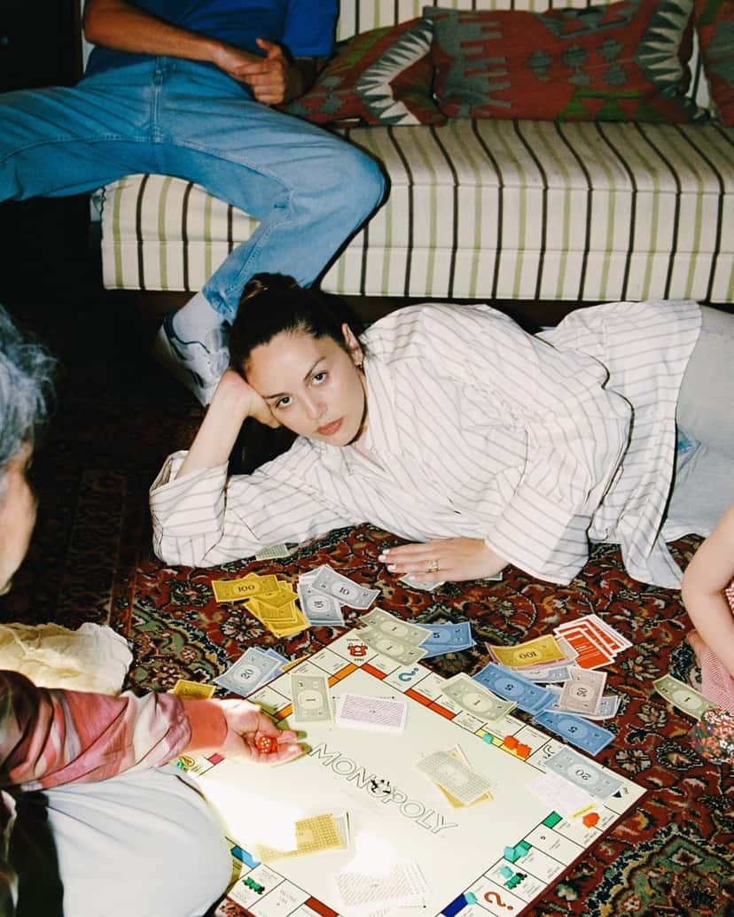 Foto de prensa de Logic1000 acostada sobre una alfombra con una camisa rayada, apoyando la cabeza en la mano, junto a un tablero de Monopoly con billetes desparramados