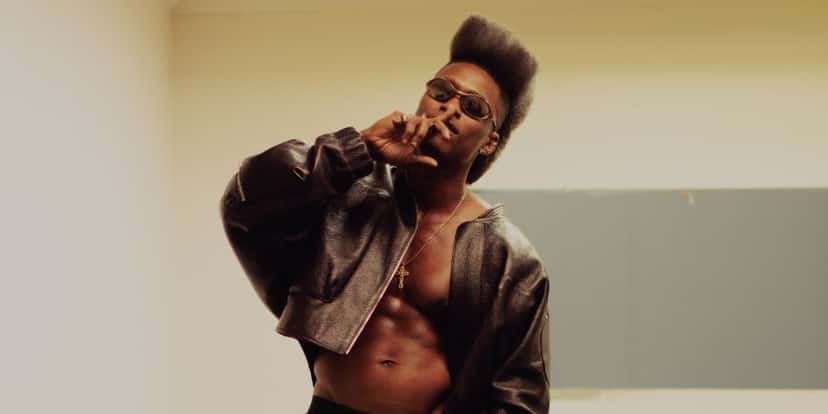 Foto de prensa de Lucky Daye con campera de cuero abierta, anteojos y peinado alto, frente a una pared clara