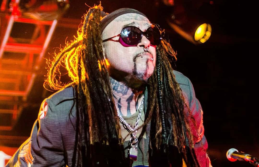 Al Jourgensen con anteojos de sol redondos, rastas y un gorro, frente al micrófono bajo luces rojas y amarillas en un show de Ministry (foto de archivo, marzo de 2015). Foto: Edifortini, CC BY-SA 4.0, vía Wikimedia Commons