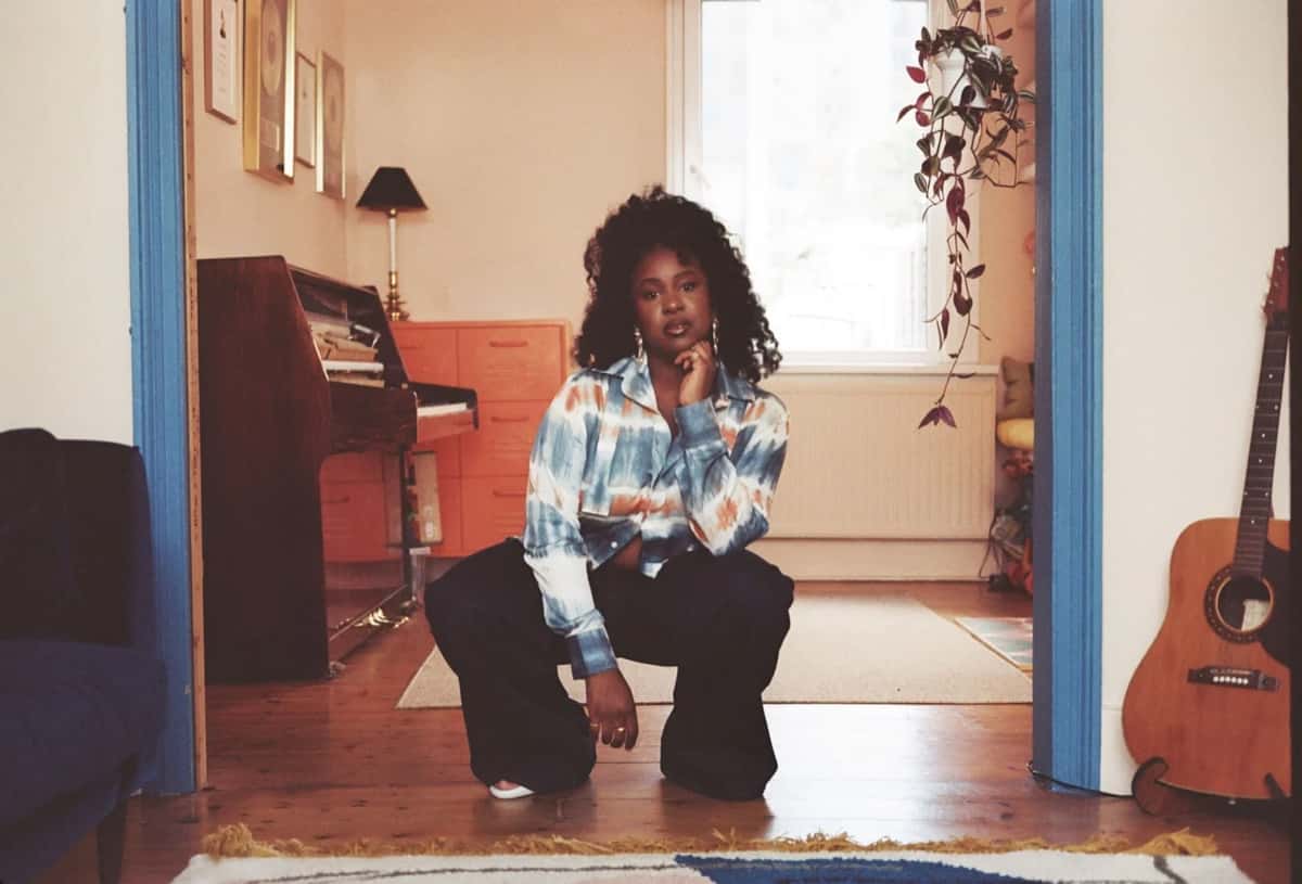 Foto de prensa oficial de Nao para "Autumn": la cantante, agachada en el umbral de una puerta de marco azul, con camisa batik celeste y naranja y pantalón oscuro ancho, apoya el mentón en la mano; detrás se ven un piano vertical, una cajonera naranja, discos enmarcados en la pared, una planta colgante y una guitarra acústica. Foto: Karolina Wielocha