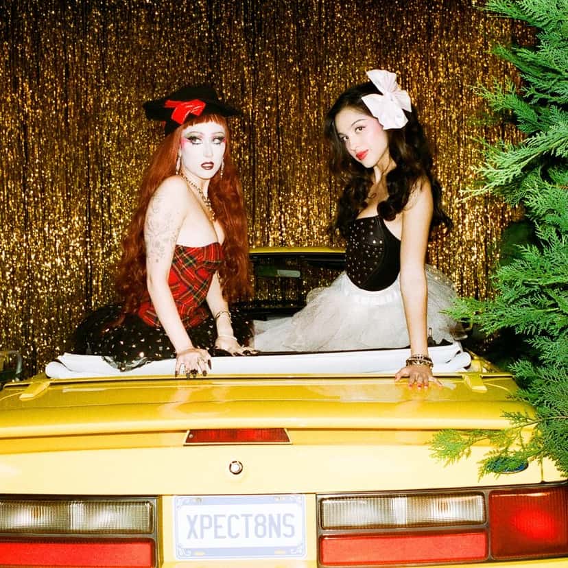 Portada de «expectations», de Olivia Rodrigo y Chappell Roan: las dos sentadas en la parte de atrás de un auto descapotable amarillo con la patente «XPECT8NS», frente a una cortina de flecos dorados