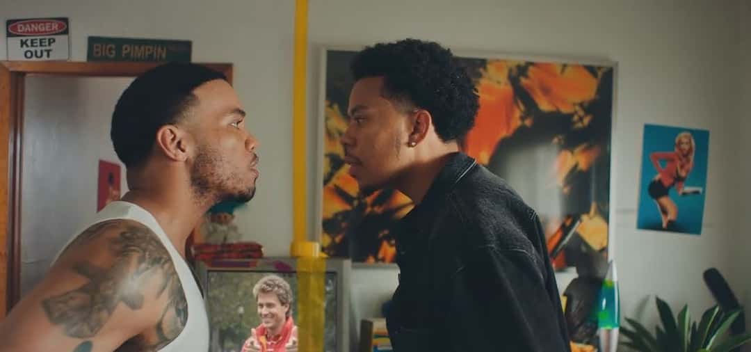 Fotograma de archivo del video oficial de «A Good Day»: el dúo cara a cara, uno con musculosa blanca y el otro con camisa negra, en un living lleno de cuadros y pósters