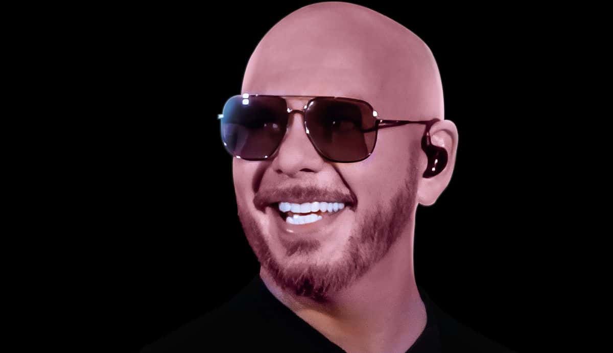 Foto de prensa oficial de Pitbull: el cantante sonríe con anteojos de sol aviador, barba candado y remera negra, sobre fondo negro