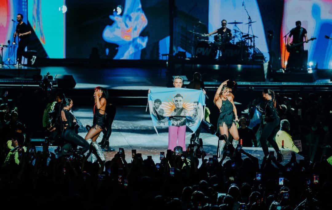 Robbie Williams con pantalón rosa sostiene una bandera argentina con su cara y el sol estampados, rodeado de bailarinas, con la banda tocando detrás y el público con celulares en primer plano