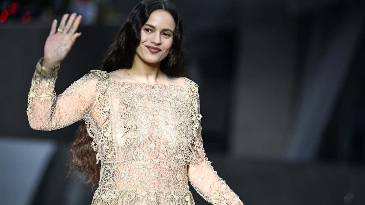 Rosalía sonriendo y saludando con la mano en alto, con el pelo largo suelto y un vestido de encaje dorado de mangas largas, sobre un fondo oscuro