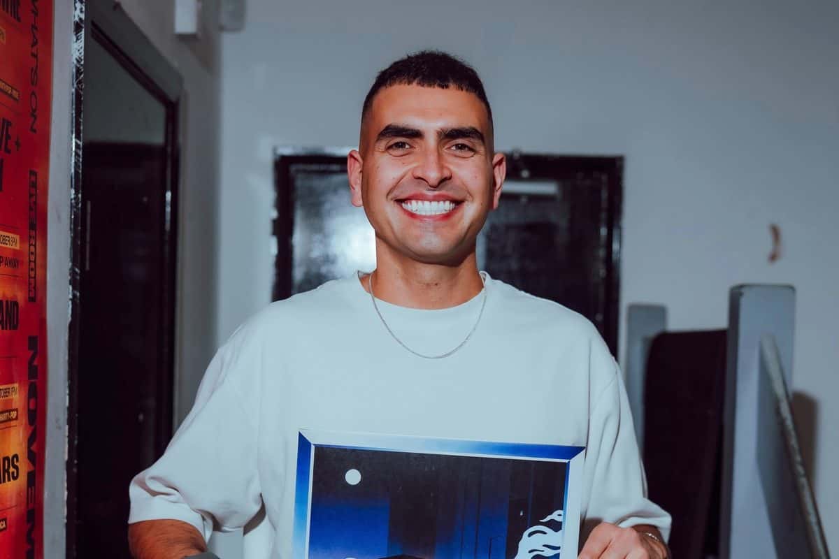 Foto de prensa de Sammy Virji: el productor inglés, sonriente, con remera clara y cadena plateada, sostiene un cuadro con la portada de «Open Late» frente a una puerta oscura