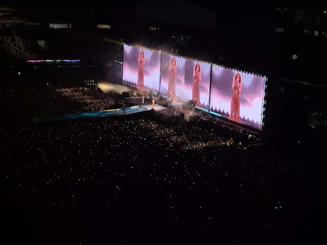 Foto de archivo de un show de la gira Las Mujeres Ya No Lloran: un estadio a oscuras con el público iluminado por celulares y una pantalla gigante que muestra a Shakira con vestido rojo, en Río de Janeiro, en 2025. Foto: Priscila Bruder, Wikimedia Commons, CC0