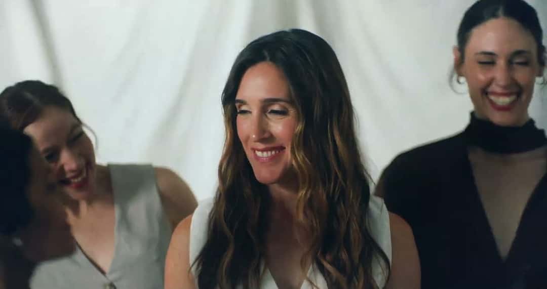 Fotograma del video oficial de «Amigas»: Soledad sonríe entre dos amigas, una con musculosa clara y otra de negro, frente a un fondo de telas blancas