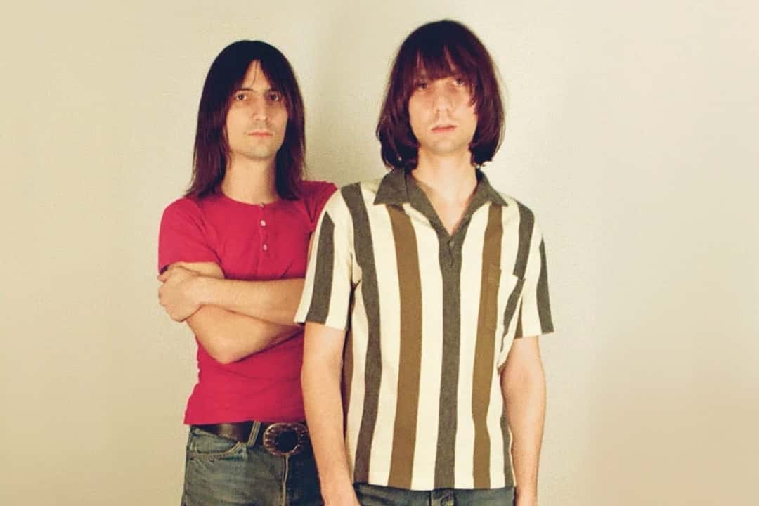 Retrato de prensa de The Lemon Twigs: los hermanos D'Addario de pelo largo sobre un fondo claro, uno con remera roja y los brazos cruzados, el otro adelante con camisa a rayas verticales marrones y blancas