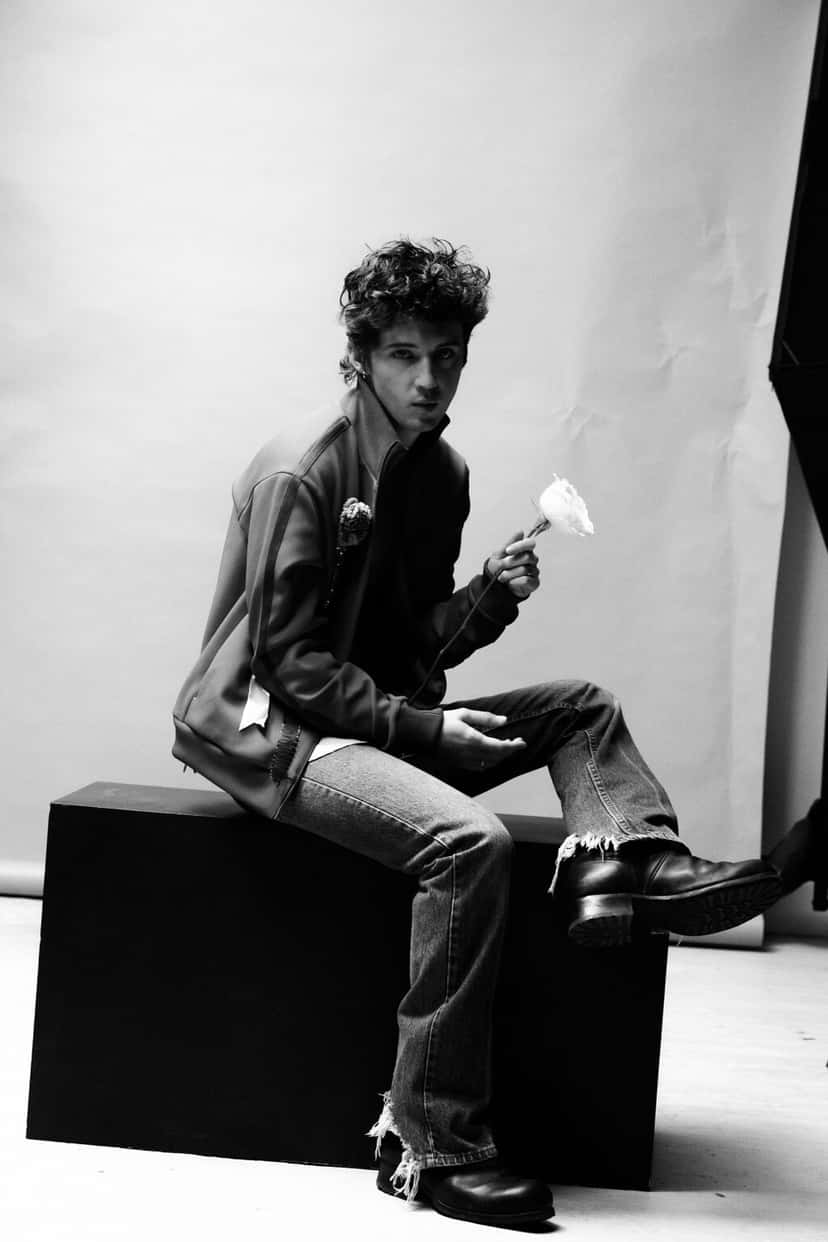 Retrato en blanco y negro de Troye Sivan sentado sobre un cubo negro, con campera deportiva, jeans gastados y una rosa blanca en la mano