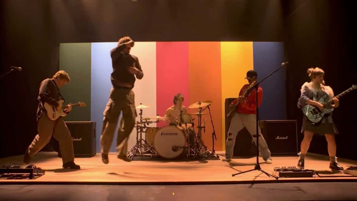 Los cinco integrantes de Turnstile tocando sobre un escenario de madera frente a un fondo de franjas verticales verde, celeste, blanca, roja, naranja, amarilla y azul; el cantante salta en el aire y el baterista toca sin remera en el centro