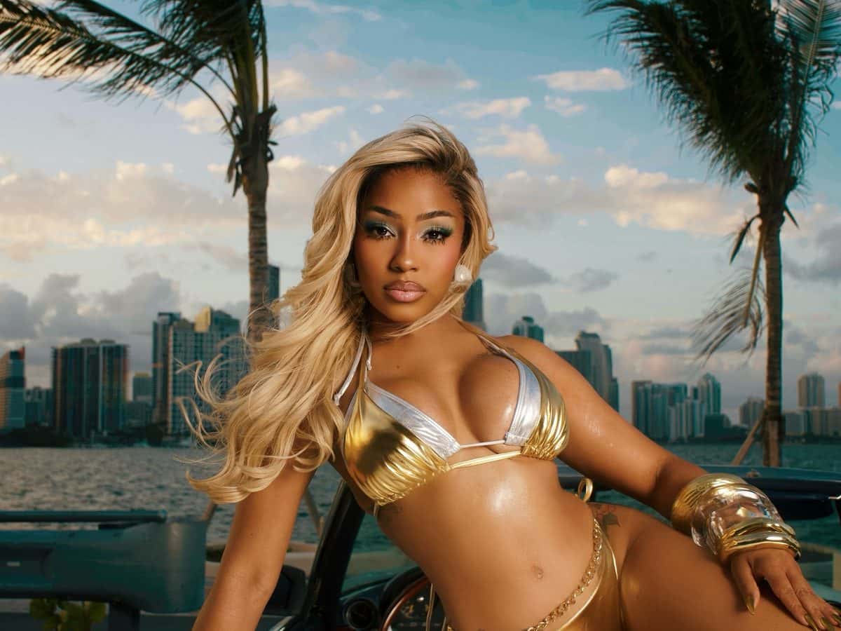 Foto de prensa de Yung Miami para «Watch Dis»: la rapera, con pelo rubio largo, bikini dorado y brazaletes, recostada sobre un auto blanco frente a la bahía de Miami, con palmeras y edificios de fondo. Foto: Brandon Almengo