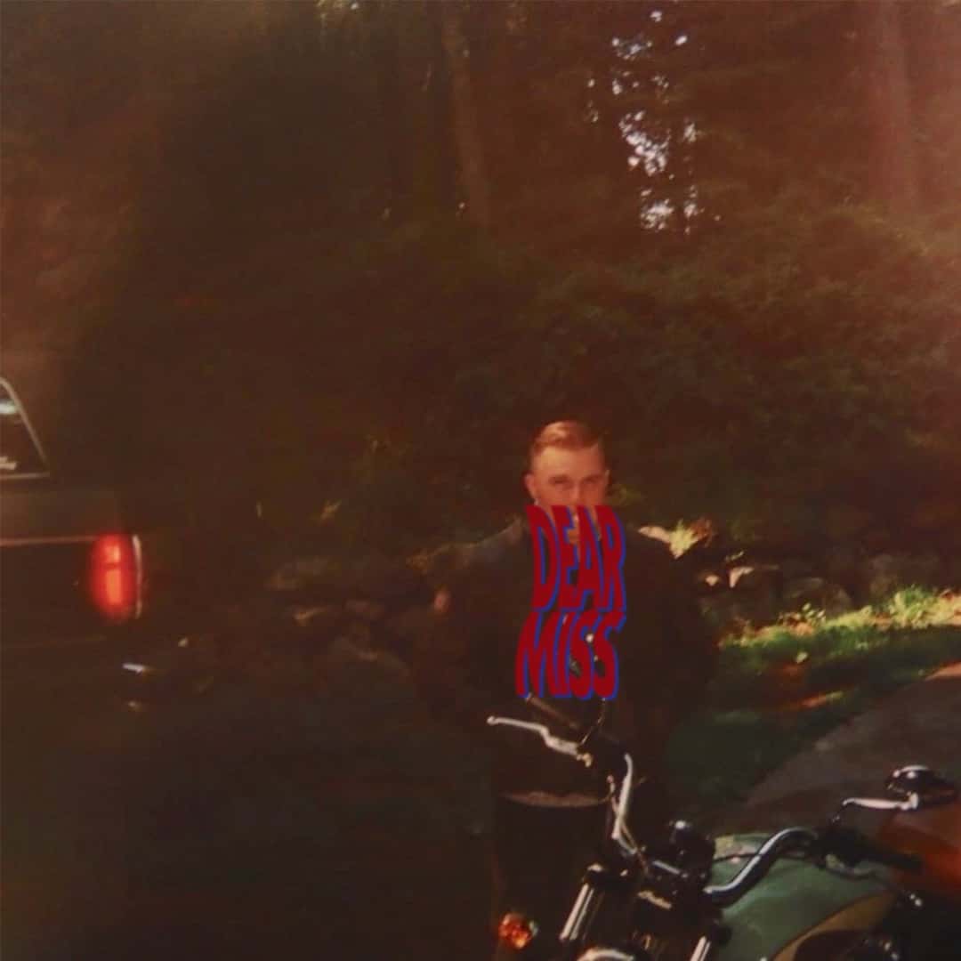 Portada del single «Dear Miss», de Zach Bryan: foto granulada de un hombre de pie junto a una moto entre árboles, con el título escrito a mano en rojo sobre su cara