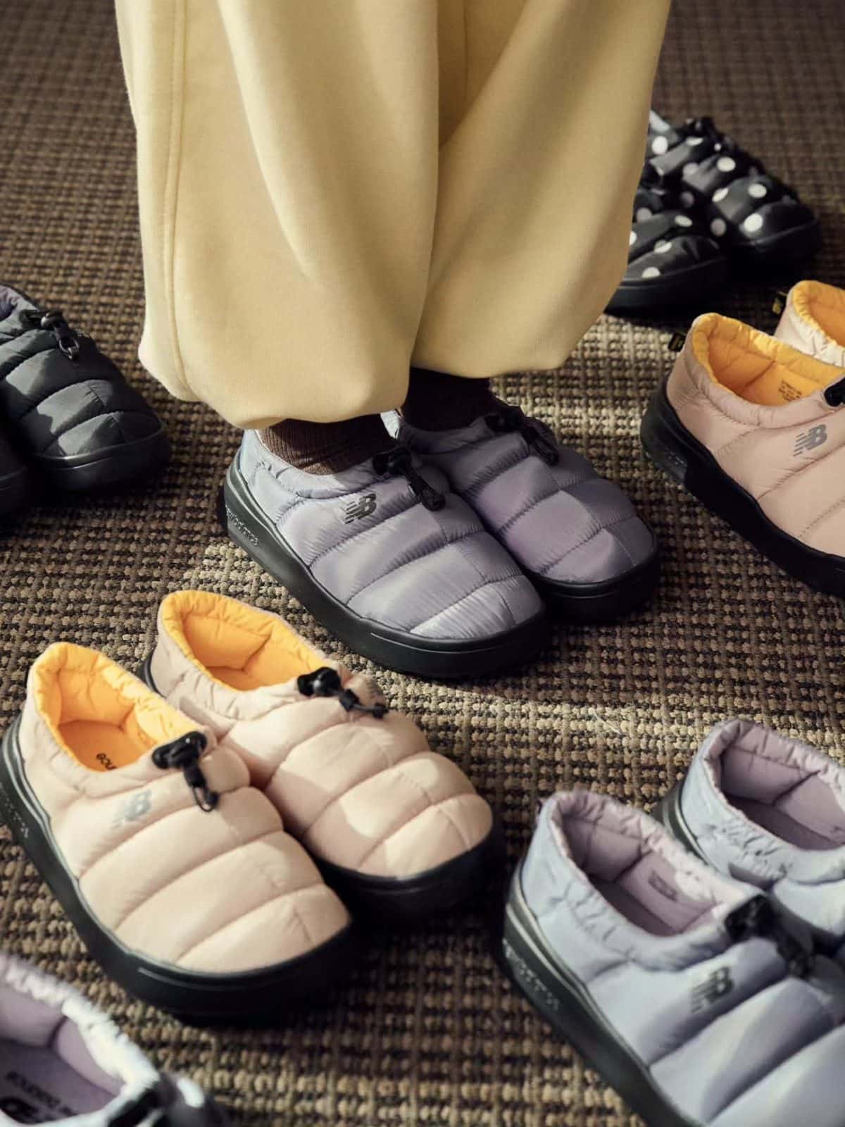 Varios pares de New Balance Sleeping Moc acolchadas en negro, gris y beige con interior amarillo, sobre una alfombra