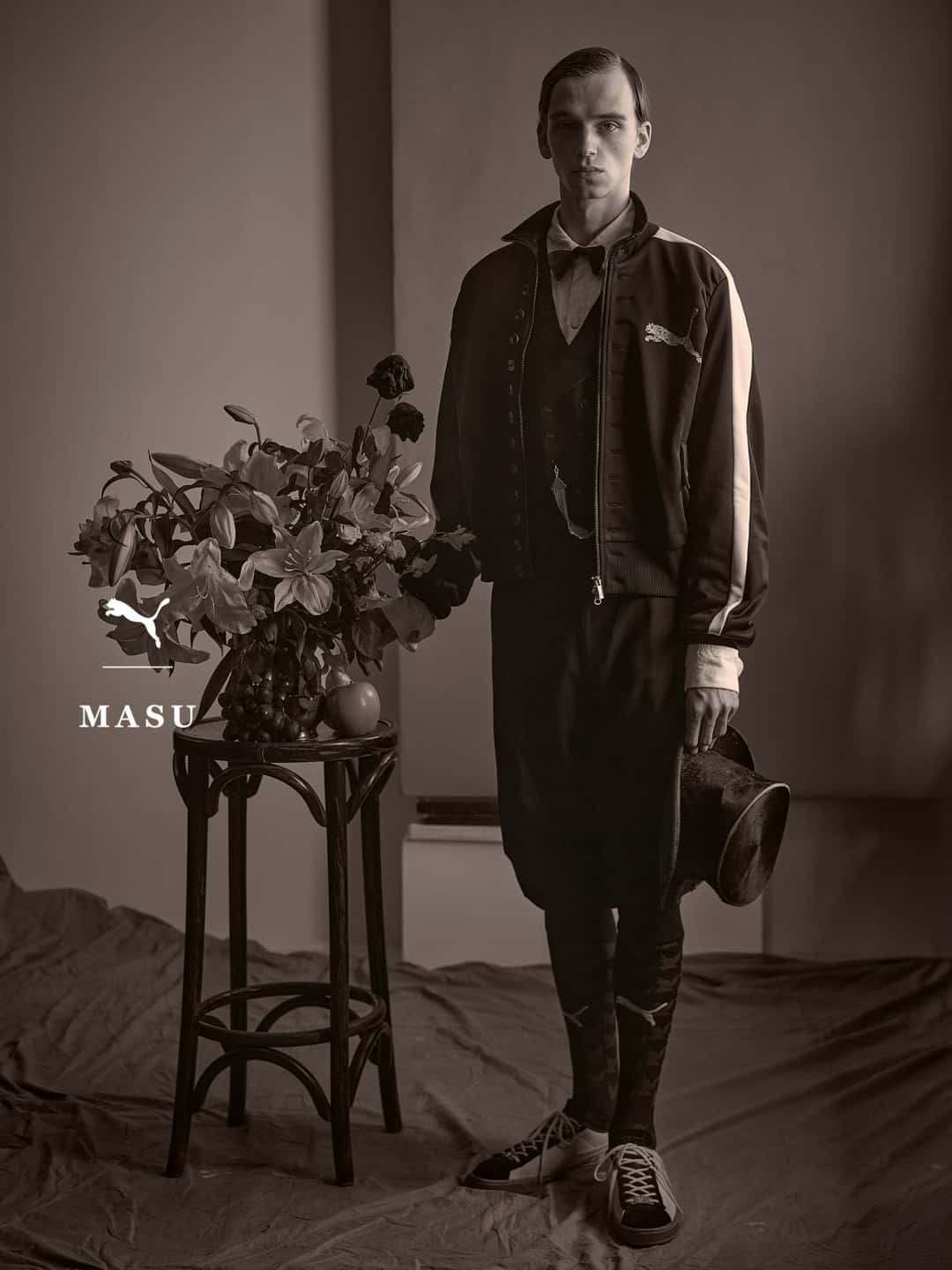 Campaña de PUMA x MASU en blanco y negro: un modelo con la campera T7 Curio negra de franjas claras, moño y sombrero en la mano, posa junto a una mesa con flores y calza las Curio Suede