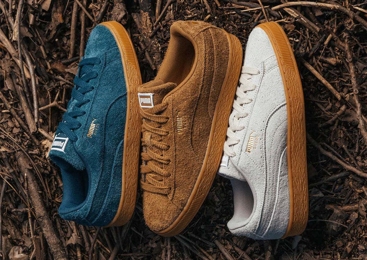 Tres PUMA Suede Forever sobre ramas secas: Midnight Petrol azul petróleo, Kona Brown marrón y Silver Fog gris claro, todos con gamuza de pelo largo, logo dorado y suela gum