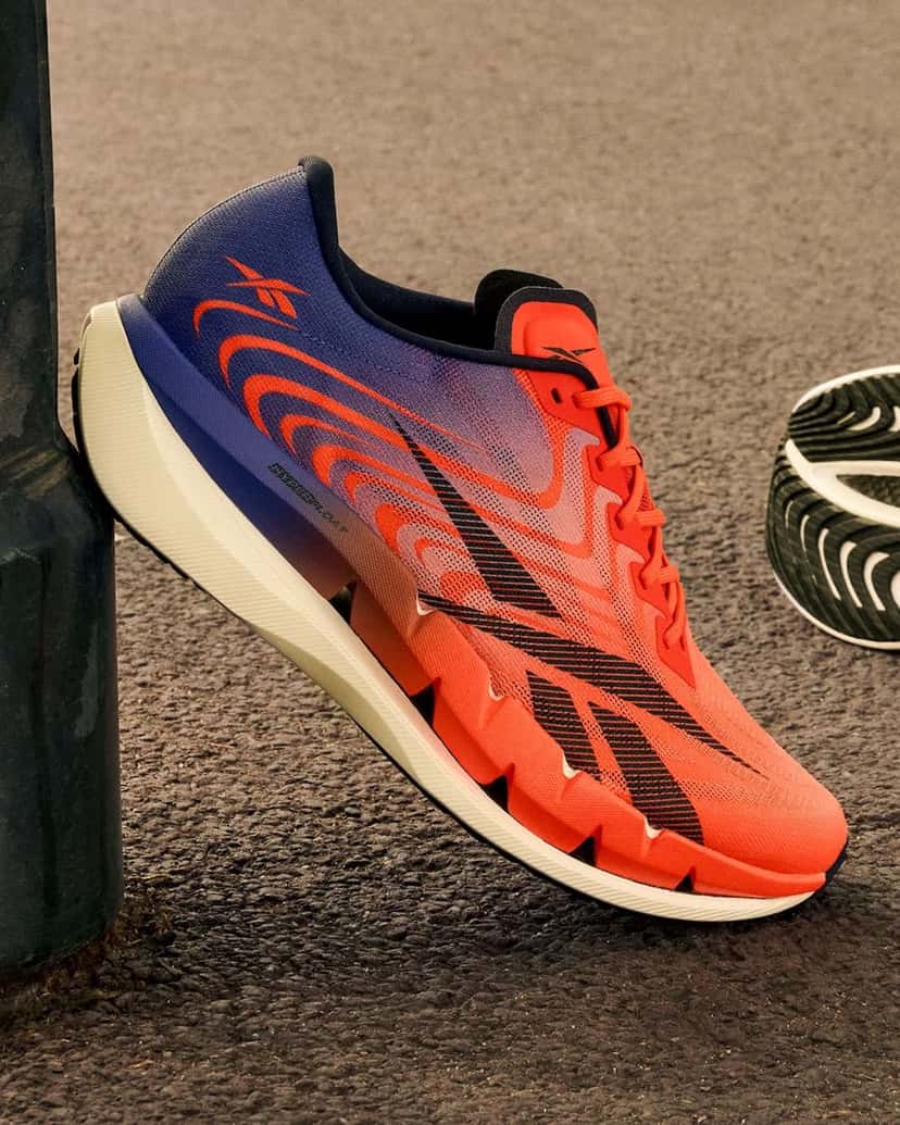 Reebok SpeedShifter apoyada contra un poste sobre el asfalto, con el upper en degradé de naranja a azul violeta y la mediasuela blanca; al costado asoma la suela de otro par
