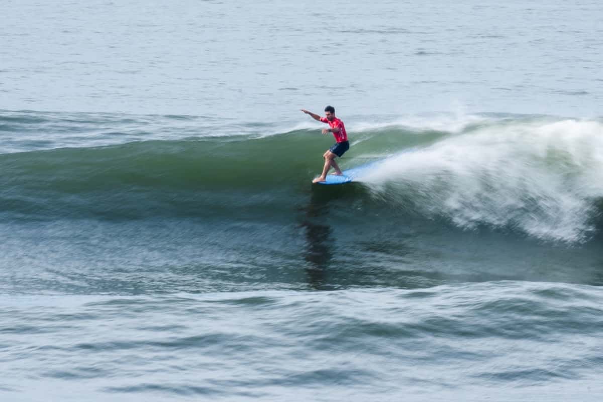Benoit «Piccolo» Clemente, con lycra roja, camina sobre un longboard celeste con un brazo extendido en una ola verde de El Sunzal