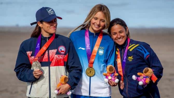 Isabella Assmann, con campera celeste y blanca de la Argentina y la medalla de oro, sonríe en el centro del podio del bodyboard femenino en Playa Grande, junto a una deportista de Chile con la plata y otra de Colombia con el bronce