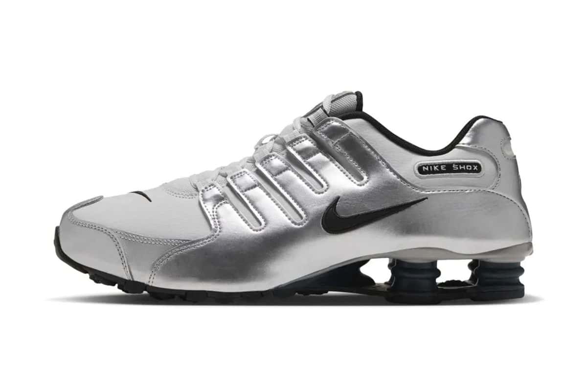Nike Shox NZ Photon Dust/Metallic Silver de perfil, con bandas plateadas brillantes, Swoosh negro y las columnas Shox del talón en gris oscuro, sobre fondo blanco